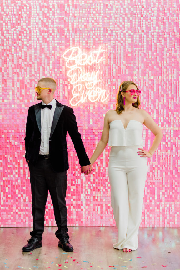a bride in a white jumper and groom in a tux in front of a pink glitter wall and a neon best day ever sign