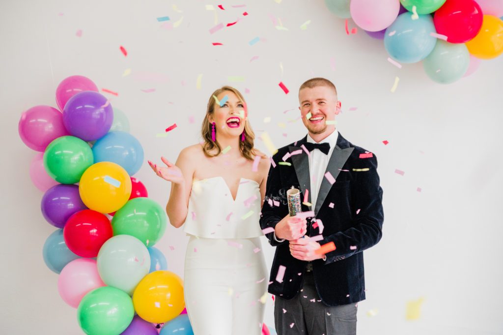 a bride in a white dress and groom in a tax popping coloful confetti in a white space with colorful balloons
